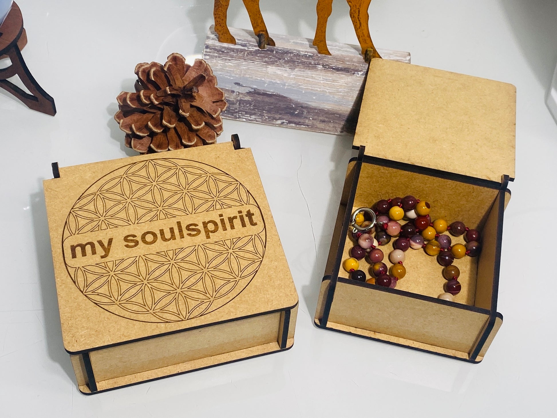 Zwei Boxen aus Holz, die linke ist geschlossen und man kann den Deckel sehen. Darauf ist eine Blume des Lebens graviert mit dem Schriftzug "mysoulspirit". Die rechte Box ist geöffnet darin liegt eine bunte Mookait Halskette mit 8 mm Perlen. 